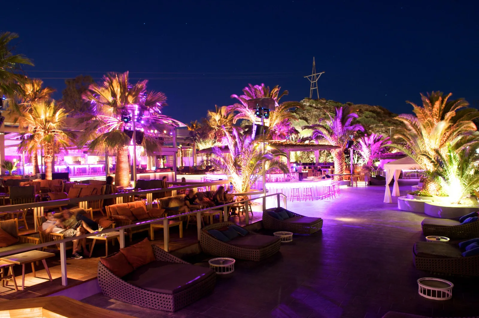 Mojito Bay seaside beach club at dusk — coloured uplighting through the palms over deck and bar, Varkiza